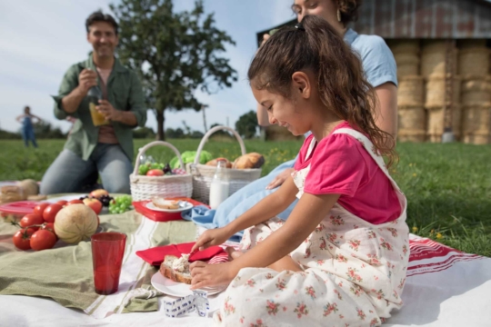Petite fille en train de pique-niquer dans l’herbe et qui tartine une tranche de pain avec du fromage fondu. Un homme et une femme sont en arrière-plan.